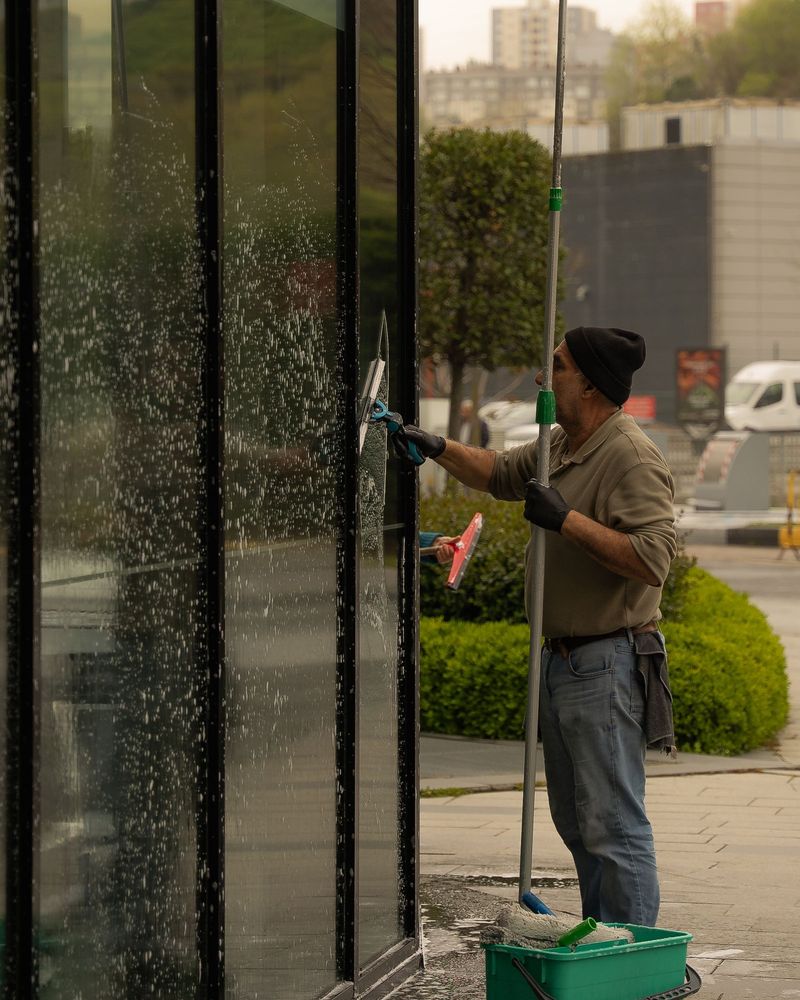 A window cleaner working a water-fed pole up a tall glass frontage, bucket at his feet. This is a stock image; a photo of Harry and the van is coming.