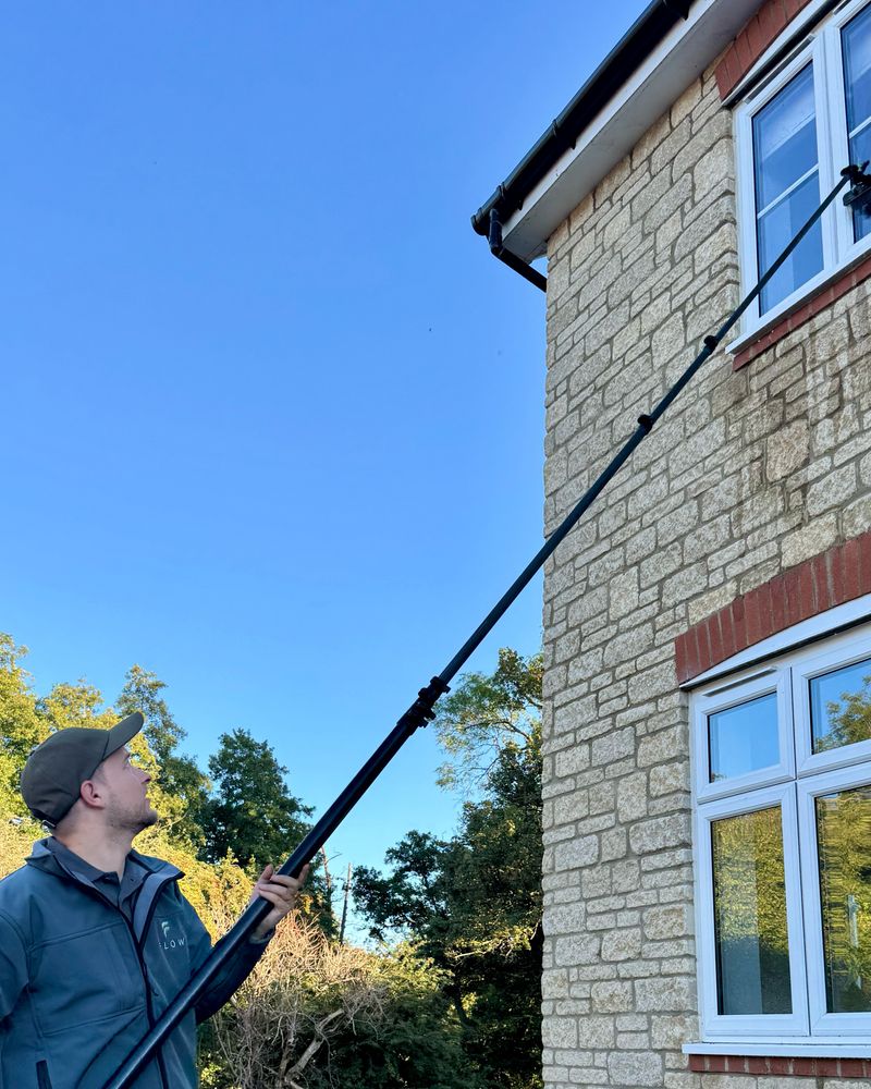 Harry reaching a first-floor window with a water-fed pole, in a Flow gilet, against a Cotswold stone house