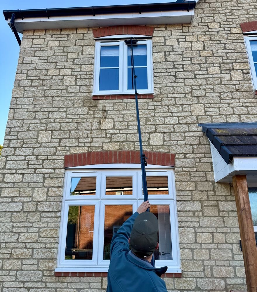 Harry reaching a first-floor window with a water-fed pole, on a Cotswold stone house under a clear sky